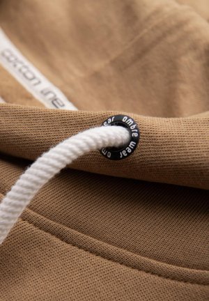 Brauner Baumwollstoff mit strukturierten Oberflächen verfügt über einen weißen Kordelzug und eine schwarze Öse mit Branding. Nahaufnahme zeigt die Nähdetails.