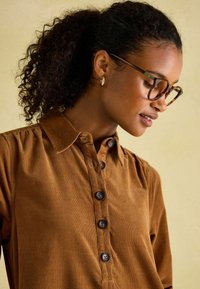 Braunes Cordhemd mit Kragen, fünf Knöpfen, leicht puffigen Ärmeln und einer glatten Textur. Das Model trägt eine Schildpattbrille.