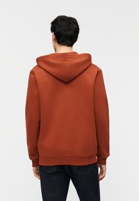 Άνδρας που φοράει φούτερ κουκούλα σκουριάς και σκούρο τζιν, στέκεται γυρισμένος πλάτη μπροστά σε απλό ανοιχτό φόντο.