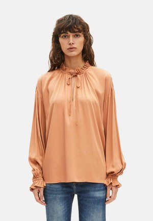 Femme aux cheveux bruns ondulés portant un chemisier satiné ample de couleur pêche avec un col et des manches à volants, associé à un jean bleu.