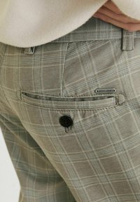Grijze geruite broek heeft een achterzak met een zwarte knoopsluiting en een merklabel. Het materiaal lijkt glad met een subtiele textuur.