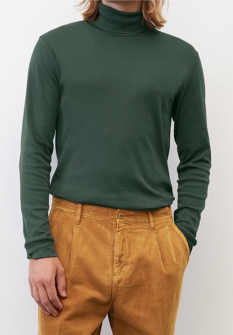 Maglietta a maniche lunghe verde con collo alto realizzata in tessuto morbido, abbinata a pantaloni in velluto a coste color senape con righe verticali e tasche frontali.