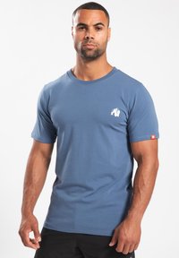 Blauwe katoenen T-shirt met korte mouwen, voorzien van een wit gorilla-logo op de borst en een oranje label op de mouw, eenvoudig ontwerp.