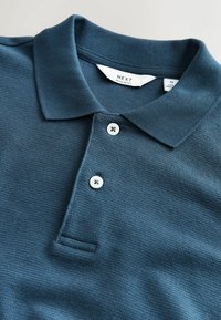 Tricou polo tricotat bleumarin cu două nasturi albi, un guler ascuțit și o etichetă vizibilă pe care scrie „NEXT Regular Fit” mărime medie.