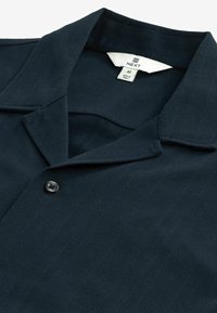 Camicetta a maniche corte blu navy scuro con colletto, chiusura con un singolo bottone e tessuto con trama. L'etichetta indica la taglia M, realizzata in Turchia.