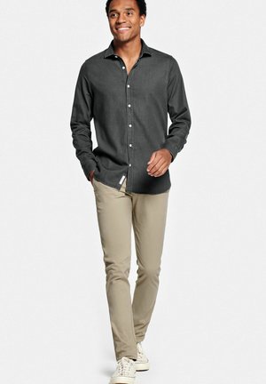 Jonge man die loopt, gekleed in een donkergrijs overhemd met knopen, beige broek en witte sneakers, glimlachend en naar de zijkant kijkend.