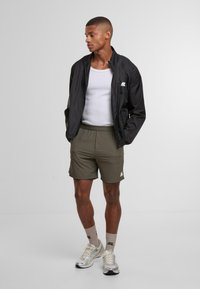 Chaqueta negra ligera sobre una camiseta sin mangas blanca, combinada con pantalones cortos verde oliva y zapatillas deportivas plateadas con calcetines beige. Detalles de logotipo simples.