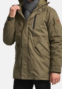 Olivgrüne isolierte Parka mit Kapuze, Reißverschluss vorne, zwei seitlichen Taschen und einer Brusttasche; aus einem glatten, strapazierfähigen Stoff gefertigt.