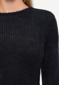 Kadar izbliza osobe koja nosi crni mekan pleteni džemper s okruglim izrezom i dugim rukavima na bijeloj pozadini.