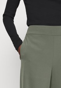 Vaatteet oliivinvihreät leveälahkeiset housut, joissa on sileä pinta, sivutaskut ja litteä vyötärö, yhdistettynä tiukkaan mustaan pitkähihaiseen puseroon.