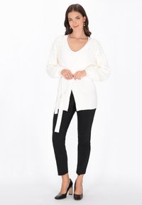 Biały kardigan z dekoltem w serek, z teksturowanymi rękawami ozdobionymi koralikami oraz przewiązaniem w talii, noszony na czarnej bluzce i spodniach.