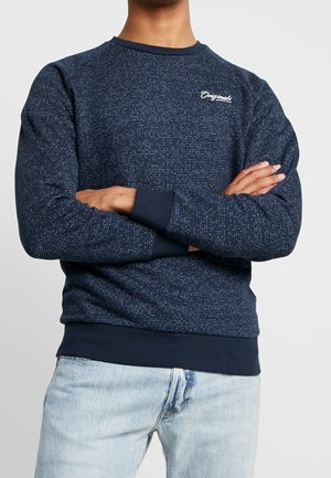 Sweatshirt bleu marine avec un motif texturé, col et poignets côtelés, et logo brodé sur la poitrine. Associé à un jeans en denim clair.