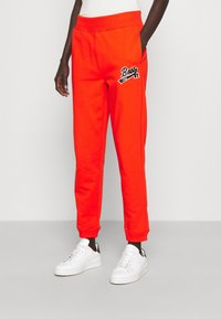 Klarröda sweatpants med ett brett elastiskt midjeband och sidofickor. Har svart broderad logotypdetalj på höften. Slät tyg.