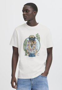 White t-shirt featuring a printed graphic of a stylized character in sunglasses and floral attire, surrounded by cacti and a circular design.