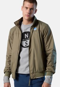 Giacca bomber verde oliva con zip sul davanti, polsini e colletto a coste; indossata sopra una felpa grigia con un logo nero.