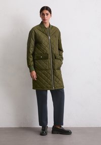 Kvinne står iført en lang olivenfarget quilted jakke, mørkeblå bukser og svarte loafers mot en ensfarget veggbakgrunn.