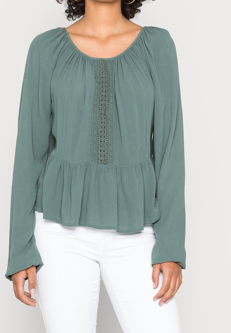 Femme portant un chemisier vert sauge à manches longues avec un détail en dentelle sur le devant et un ourlet peplum, associé à un pantalon blanc.