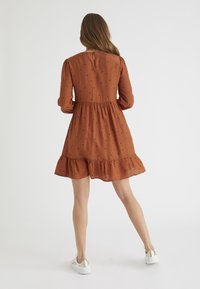 Vestido de color óxido con mangas largas, cintura fruncida y dobladillo con volantes. Presenta lunares negros y un cierre de botones en la parte posterior.