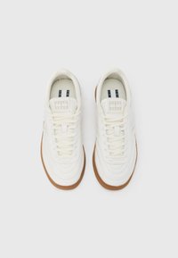 Λευκά δερμάτινα αθλητικά παπούτσια με υφή στην επιφάνεια, στρογγυλεμένη μύτη και εξωτερική σόλα από καουτσούκ χρώματος καραμέλας. Διαθέτουν λευκά κορδόνια και ετικέτα με λογότυπο στη γλώσσα.