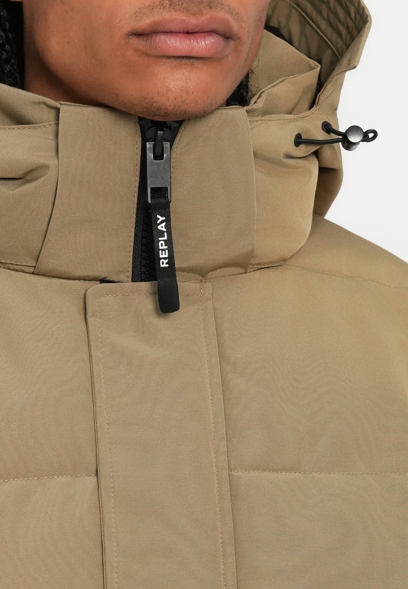 Beige Steppjacke mit hohem Kragen, Reißverschluss vorne und sichtbarem Markenetikett. Verfügt über vertikale Nähte und verstellbare Kapuzenkordeln.