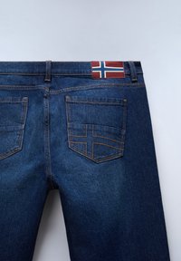 Vista posteriore di jeans in denim blu scuro con cuciture arancioni e una patch della bandiera norvegese sopra la tasca destra.