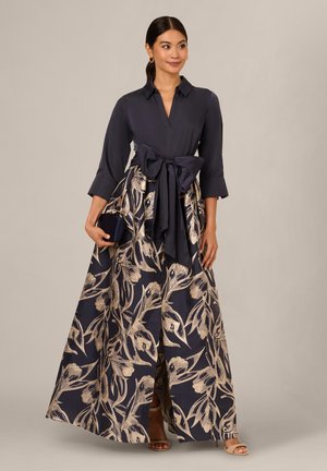 Kvinde iført en navyblå langærmet kjole med stort bælte med sløjfe i taljen og nederdel med gyldent blomsterprint, som holder en navy clutch og beige hæle.