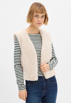 Beige Kunstfellweste mit offenem Vorderteil und breitem Kragen, getragen über einem schwarz-weißen gestreiften Langarmshirt und blauen Jeans.