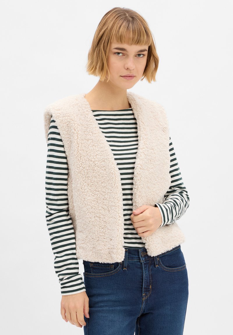 Beige nepbont vest met een open voorkant en brede kraag, gedragen over een zwart-witte gestreepte langemouwen shirt en blauwe jeans.