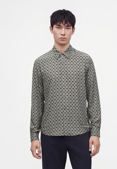 Jeune homme portant une chemise grise à motifs avec des boutons et des manches longues, ainsi qu'un pantalon foncé, debout devant un fond clair uni.