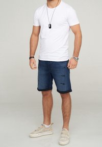 Camiseta blanca de algodón, pantalones cortos de mezclilla azul con dobladillo deshilachado y zapatos de color beige claro. Los accesorios incluyen un collar de placa negra y un reloj de pulsera.