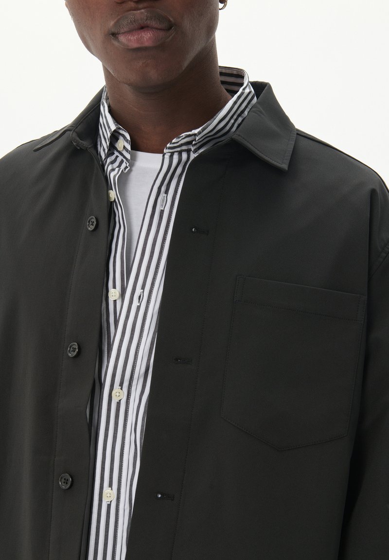 Czarna koszula z zapięciem na guziki o teksturowanym wykończeniu, nałożona na białą i czarną koszulę w paski, z kieszenią na piersi i widocznymi guzikami.