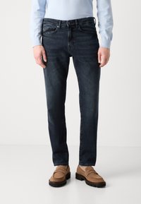 Hombre vistiendo jeans slim-fit azul oscuro, suéter azul claro y mocasines de ante color beige de pie contra un fondo blanco liso.