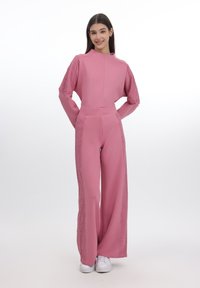 Rosa jumpsuit med blondekanter, med høy hals, brede ermer og løstsittende bukser, kombinert med hvite joggesko.