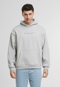 Világosszürke kapucnis pulcsi puha anyagból, elülső zsebbel és húzózsinóros kapucnival. A mellkason a "JUST ANOTHER HOODY" felirat van belegravírozva.