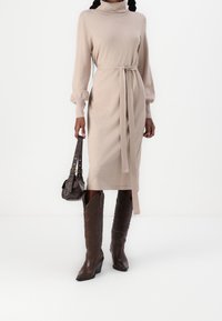 Vestito beige in maglia con collo alto, maniche lunghe e cintura annodata. Abbinato a stivali da cowboy in pelle marrone e una piccola borsa marrone.