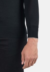 Maglione nero a maglia con orlo e polsini a costine. I polsini presentano cuciture argentate a contrasto. Mostra il braccio e la mano su uno sfondo neutro.