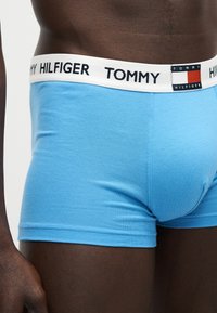 Άνδρας κορμός που φοράει ανοιχτό μπλε boxer Tommy Hilfiger με λευκή ζώνη μέσης και ορατό λογότυπο της μάρκας.