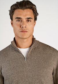 Brązowy sweter z zamkiem błyskawicznym wykonany z miękkiego materiału dzianinowego, z wysokim kołnierzem, nałożony na biały T-shirt. Zbliżenie, neutralne tło.