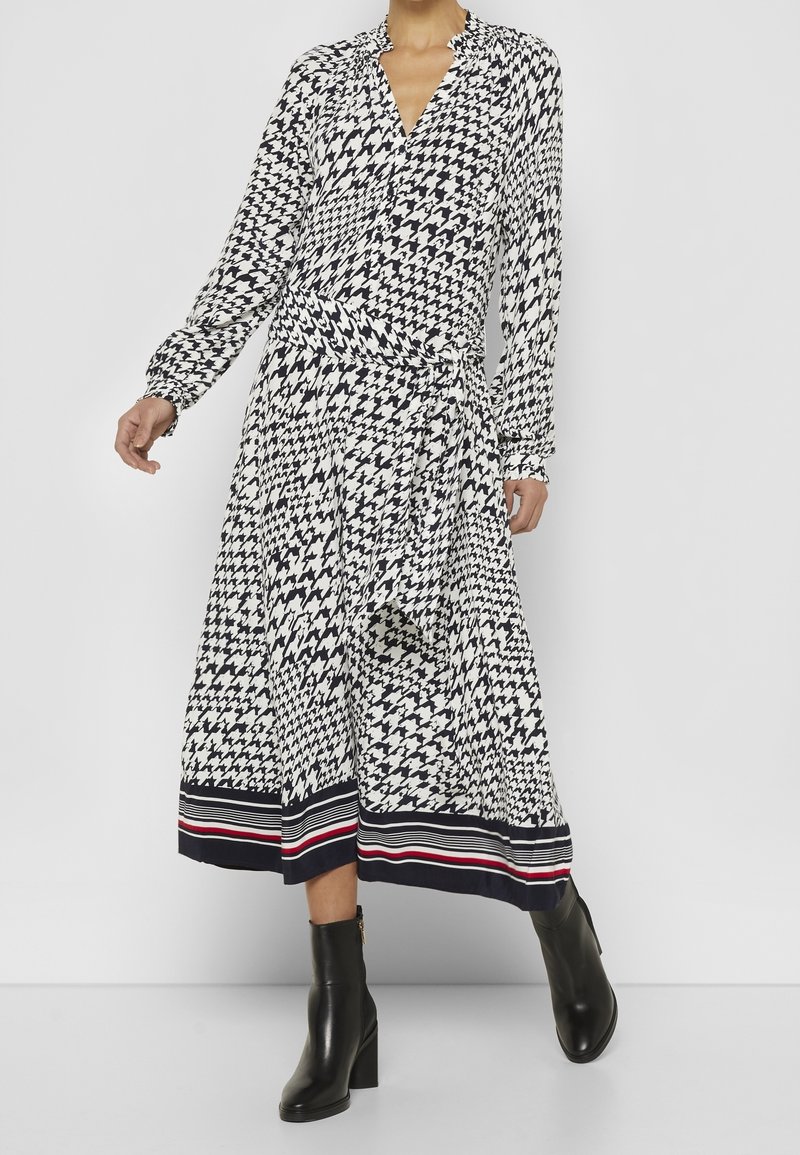 Långärmad omlottklänning i svart och vit hounds-tooth-mönster, med en djup v-ringning och en randig nederdel i marinblått, rött och vitt.