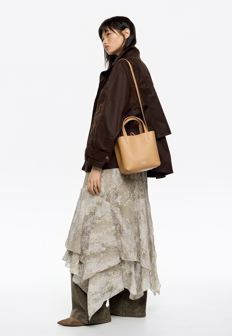 Borsa tote in pelle marrone con manici circolari, indossata sopra un vestito grigio chiaro a strati e un giubbotto marrone. Materiali texturizzati presenti.