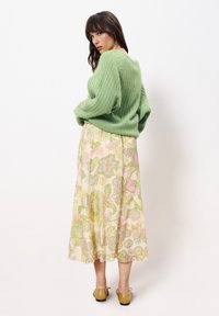 Grüner, gerippter Pullover kombiniert mit einem langen, gemusterten Rock mit floralen Designs in sanften Farben. Getragen mit hellgoldenen Schuhen.