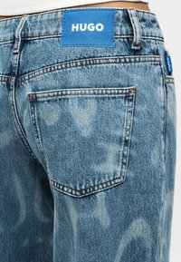 Ξεθωριασμένο μπλε τζιν με ετικέτα στο πίσω μέρος που γράφει "HUGO", εμφανή ραφές και σχέδιο με τσεπάκι.