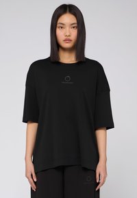 Svart kortärmad T-shirt med en lös passform, med rund halsringning och en subtil logotypdetalj i mitten. Slät textur och minimalistisk design.