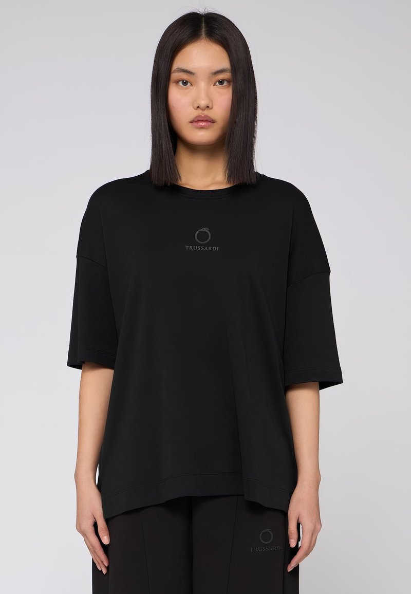 Svart kortärmad T-shirt med en lös passform, med rund halsringning och en subtil logotypdetalj i mitten. Slät textur och minimalistisk design.