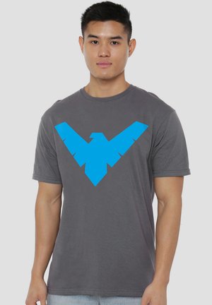 Young man stands facing forward wearing a gray t-shirt with a large stylized blue bird logo on the chest against a plain background.