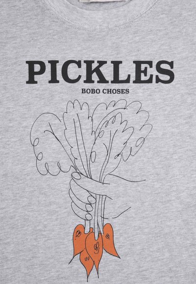Hand hält einen Bund Blattgemüse mit orangefarbenen Wurzeln, darüber der Text "PICKLES" und "BOBO CHOSES" auf grauem Stoff.