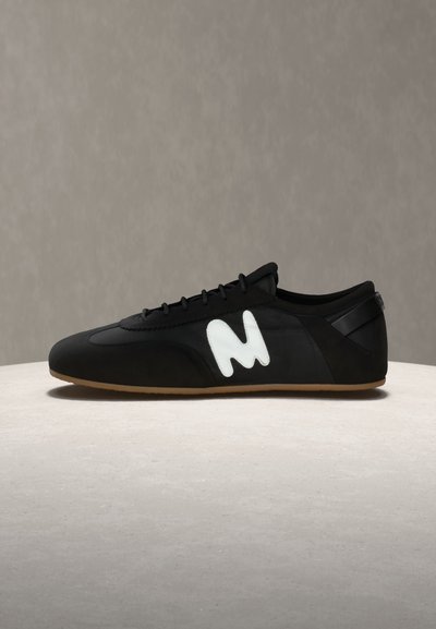 Μαύρο αθλητικό παπούτσι χαμηλού προφίλ με καφέ σόλα και λογότυπο "M" σε λευκό στυλιζαρισμένο σχέδιο στο πλάι, τοποθετημένο σε ανοιχτό γκρι επιφάνεια με γκρι φόντο.
