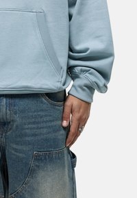 Sudadera azul claro con un bolsillo tipo canguro, puños acanalados, combinada con jeans azules desgastados que cuentan con un bolsillo lateral y un anillo plateado.