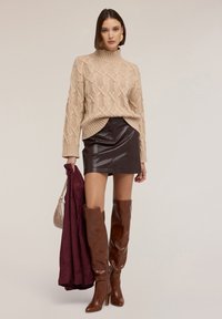 Beżowy sweter dziewiarski z wysokim kołnierzem i wzorami w warkocze, zestawiony z brązową skórzaną mini spódnicą i wysokimi brązowymi kozaczkami, trzymający bordową kurtkę.