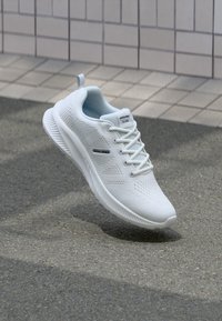 Weißer leichter Sportschuh mit Mesh-Design und Schnürsenkeln, schwebt über Asphalt in der Nähe einer gefliesten Wand.